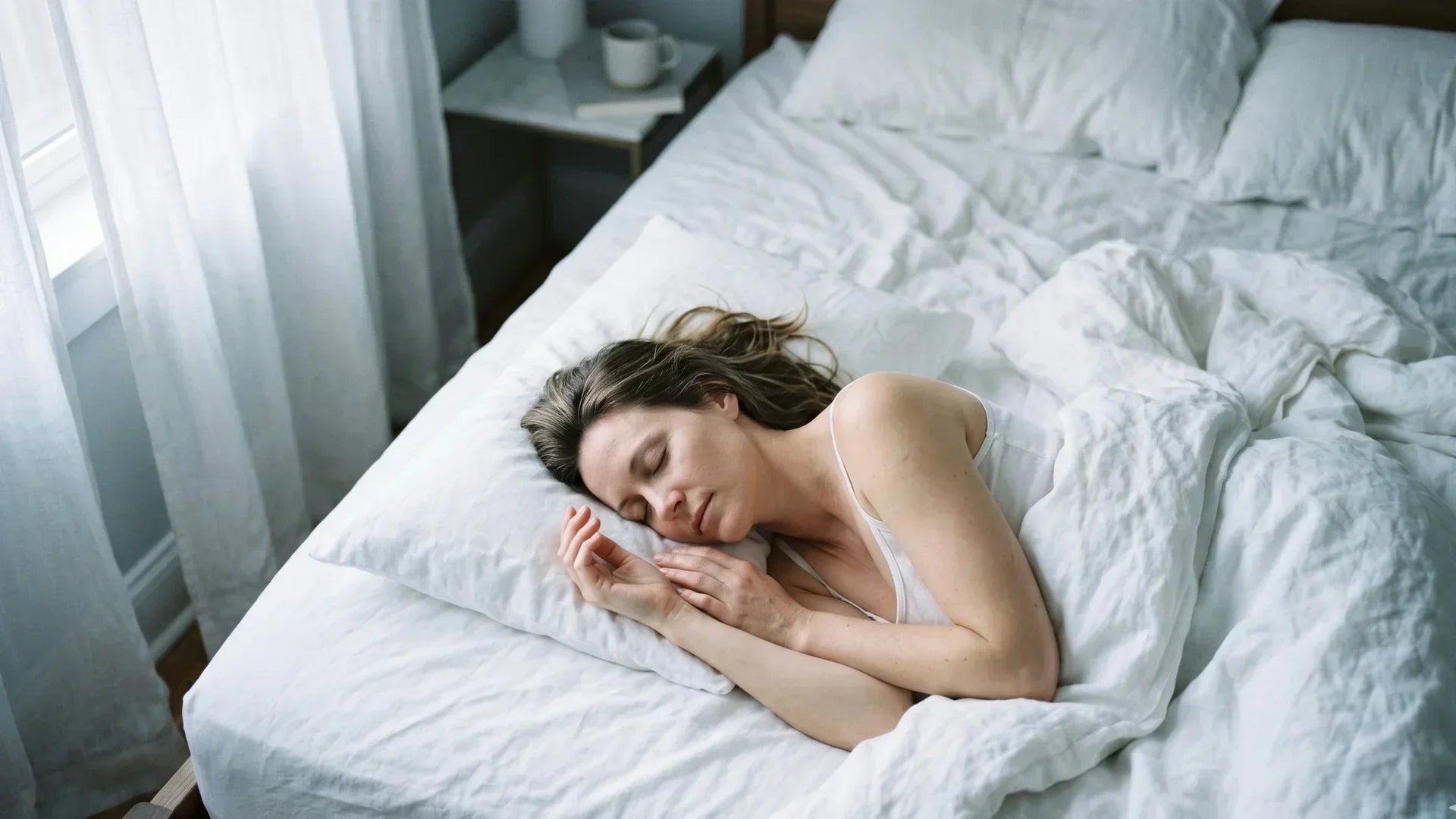 Woman in her late 30s resting peacefully on white linen bedding in soft cool morning light