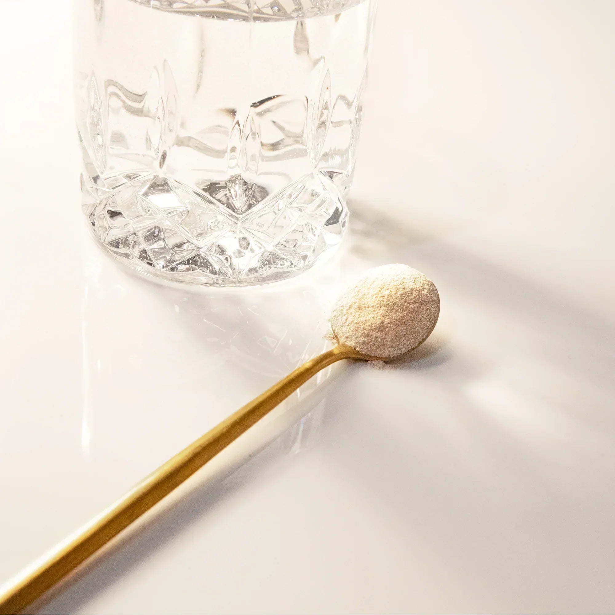 A gold spoon with a scoop of collagen powder resting beside a glass of water on a clean white surface